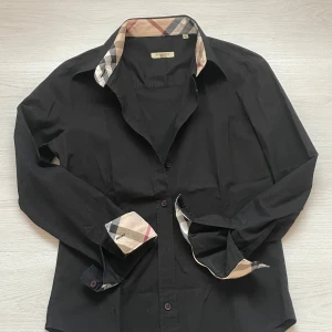 Svart skjorta från Burberry med rutiga detaljer - Säljer en svart skjorta från Burberry med klassiska beige, röda och svarta rutiga detaljer på krage och ärmslut. Skjortan har lång ärm och knäppning framtill. Perfekt för dig som gillar stilrena plagg med en twist. Står M men är S