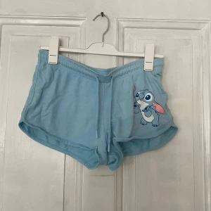 Ljusblå Disney shorts med Stitch-tryck - Säljer ett par ljusblå shorts från Disney med ett gulligt Stitch-tryck på ena benet. Shortsen har elastisk midja med snörning och är tillverkade i mjuk bomull. Perfekta för sommaren eller som mysplagg hemma.
