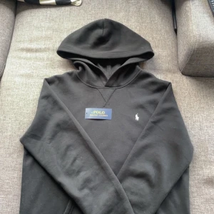 Svart hoodie från Polo Ralph Lauren - Säljer en klassisk svart hoodie från Polo Ralph Lauren med liten broderad logga på bröstet. Tröjan har huva och är tillverkad i mjukt material, perfekt för en avslappnad stil.