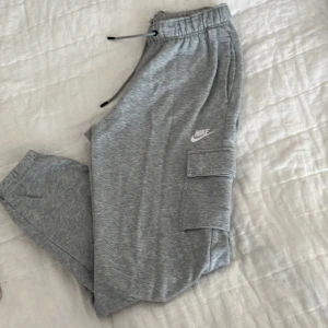 Grå mjukis cargobyxor från Nike - Säljer ett par grå mjukis/cargobyxor från Nike med resår och dragsko i midjan. Byxorna har stora fickor på sidorna och Nike-logga tryckt på ena benet. Perfekta för en avslappnad och sportig stil.