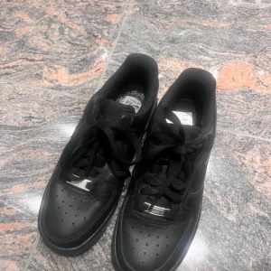 Svarta Nike Air Force 1 sneakers - Säljer ett par klassiska svarta Nike Air Force 1 sneakers i helsvart läder med svart sula och snörning. Ikonisk Nike-logga på sidan och AIR-detalj på hälen. Perfekta för dig som gillar stilrena och tidlösa sneakers. Storlek 35,5 och det går att snacka om priset :)