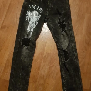 Svarta jeansbyxor med ängeltryck från Amiri - Säljer ett par svarta jeansbyxor från Amiri med coolt ängeltryck och stora slitningar på benen. Byxorna har en tvättad look, raka ben och klassiska bakfickor. Perfekta för dig som vill sticka ut med din stil.