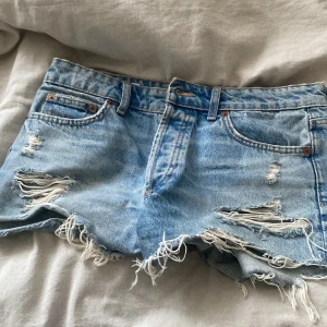 Blå jeansshorts med fransar från Zara - Säljer ett par ljusblå jeansshorts från Zara med slitna detaljer och fransig kant. Shortsen har klassisk femficksdesign, knappar i gylfen och låg midja. Perfekta för sommaren och ger en cool, avslappnad look.