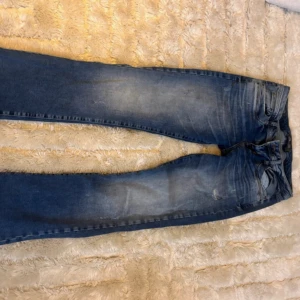 Blå jeans med slitningar - Säljer ett par klassiska blå jeans med lätt slitna detaljer och normal passform. Jeansen har fem fickor och stängs med dragkedja och knapp. Perfekta till vardags och ger en avslappnad look.