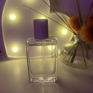 Ariana Grande parfym med lila kork - Säljer en stilren parfym från Ariana Grande i en genomskinlig, fyrkantig glasflaska med lila kork. Flaskan har minimalistisk design och texten 'Ariana Grande' tryckt på sidan. Ej kartong