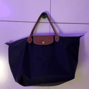 Svart Longchamp Le Pliage handväska - Klassisk svart Le Pliage handväska från Longchamp med bruna skinnhandtag och lock. Väskan har guldfärgad knapp med logga och dragkedja med Longchamp-märke. Perfekt storlek för att få plats med allt du behöver och stilren design. Storlek M.