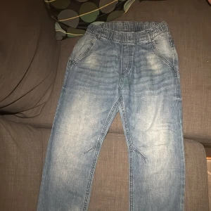 Blå jeansbyxor med resårmidja - Säljer ett par ljusblå jeansbyxor med resår i midjan och avslappnad passform. Byxorna har klassiska fickor fram och bak, samt snygga slitningar för en cool look. Perfekta för dig som gillar bekväma och stilrena jeans.