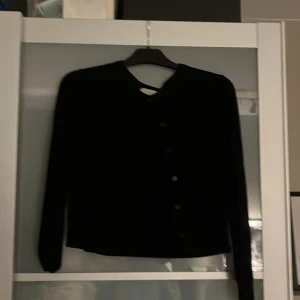 Svart kofta från H&M, storlek M - En svart, klassisk kofta från H&M i storlek M. Koftan har långa ärmar, rund halsringning och knäppning framtill med svarta knappar. Perfekt att slänga över en t-shirt eller linne för en enkel och stilren look.