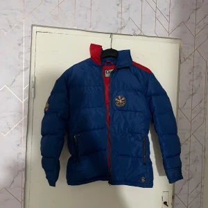 Blå och röd pufferjacka Campri S - Retro pufferjacka från Campri i blått med röda detaljer och broderad Cervinia-patch på bröstet. Jackan har hög krage, dragkedja framtill och två fickor med dragkedja. Perfekt för kalla dagar när du vill sticka ut med färg.