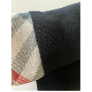 Svart ulltröja Burberry med rutiga detaljer - Svart långärmad tröja från Burberry i 100% ull. Klassisk passform med ribbade muddar och ikoniska rutiga detaljer i beige, rött och grått på ärmsluten. Perfekt för dig som gillar stilrena plagg med en twist från ett lyxigt märke.