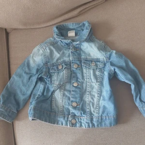 Ljusblå jeansjacka från H&M, strl 86 - Snygg ljusblå jeansjacka från H&M i klassisk modell med knappar framtill och två bröstfickor. Jackan har långa ärmar och coola sömdetaljer. Perfekt för dig som vill ha en tidlös och trendig look till de små.