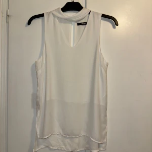 Vit ärmlös blus från BikBok - Säljer en stilren vit blus från BikBok i storlek S. Blusen är ärmlös med klassisk krage och en liten slits framtill. Den har en lätt, flowy känsla och är tillverkad i ett tunt, mjukt material som faller snyggt. Perfekt att styla med jeans eller kjol.