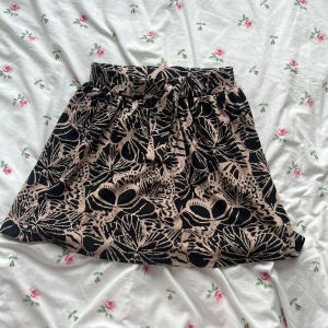 Svart och beige kjol från Pomp de Lux - Supersnygg kortkjol från Pomp de Lux i svart och beige med ett coolt blommigt mönster. Kjolen har bred resår i midjan och en söt knyt-detalj framtill.