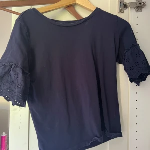 Mörkblå blus med spetsärm KappAhl - Supersöt mörkblå blus från KappAhl med korta ärmar i broderad spets och lätt volang. Topp i mjuk bomull med rund halsringning och relaxed fit. Perfekt för dig som gillar stilrena detaljer och en feminin touch. Inga hål.