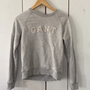Grå sweatshirt från GANT - Grå sweatshirt från GANT med broderad logga framtill. Klassisk rund halsringning, ribbade muddar och lång ärm. Perfekt för en chill och stilren look. Passar dig som gillar enkel och tidlös stil.