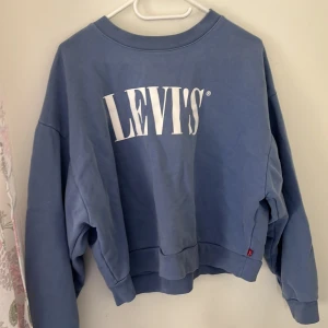 Blå sweatshirt från Levi's med logga - Blå sweatshirt från Levi's i bomull med stor vit logga framtill. Tröjan har en loose passform, rund halsringning och långa ärmar. Perfekt för en chill och avslappnad stil. Klassisk och enkel design som funkar till det mesta.
