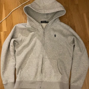 Grå hoodie från Polo Ralph Lauren - Snygg ljusgrå hoodie från Polo Ralph Lauren med dragkedja och klassisk huva. Tröjan har en broderad logga på bröstet, känguruficka framtill och är tillverkad i mjuk bomullsmix. Perfekt för chill dagar eller när du vill ha en clean streetstyle.