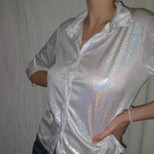 Glittrig silver skjorta med trekvartsärm - Säljer en glittrig silverfärgad skjorta med trekvartsärm och klassisk krage. Skjortan har knappar framtill och är tillverkad i ett syntetmaterial som ger en skimrande effekt. Perfekt för dig som vill sticka ut med en unik och trendig look! Passar både S och M