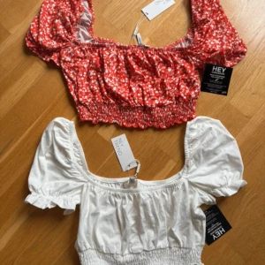 2 puffärmade croppade toppar Nelly - Aldrig använda💗Två söta croppade toppar från Nelly med puffärmar och smockad midja. En är vit och den andra är röd med små vita blommor. Båda har fyrkantig urringning och volangdetaljer vid ärmslut. Perfekta för en trendig och somrig look!