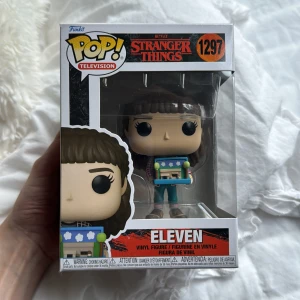 Stranger Things: Eleven (Funko Pop! #1297) - Samlarfigur av Eleven från Netflix-serien Stranger Things. Perfekt för fans av popkultur och spännande berättelser! Eleven håller en färgglad låda och har sin ikoniska look. En cool detalj till din hylla eller present till någon som älskar serien.