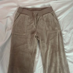 Beige velourbyxor Juicy Couture XS - Supermjuka beige velourbyxor från Juicy Couture med bred resår i midjan och snörning. Byxorna har raka ben, stora fickor fram och bakfickor med knapp. Perfekta för en chill och trendig look. Nypris 1200 kr.