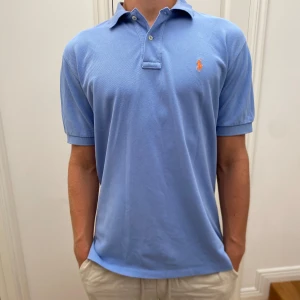 Ralph Lauren piké - Snygg ljusblå pikétröja från Ralph Lauren med klassisk krage och två knappar. Tröjan har korta ärmar och den ikoniska orangea polospelaren broderad på bröstet. Tillverkad i mjuk bomull som känns skön mot huden. Perfekt för en clean och stilren look på stranden. Modellen är 184 cm. 