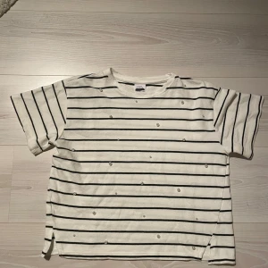 Randig t-shirt med pärlor från Zara - Vit och svart randig t-shirt från Zara med pärldetaljer på framsidan. Tröjan har korta ärmar, rak passform och små slitsar i sidorna. Materialet är mjukt och känns som bomull. 
