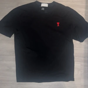 Svart t-shirt med rött hjärta Ami - Svart t-shirt från AMI med klassisk passform och ett litet rött hjärta broderat på bröstet. Enkel och stilren design som passar perfekt till jeans eller shorts. Mjuk bomullskvalitet och rund halsringning.