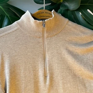 stickad half zip-tröja - Snygg beige stickad tröja från Oscar Reiner med hög krage och half zip-dragkedja. Tröjan har lång ärm och en clean, stilren look som passar perfekt till lager-på-lager. Ribbad detalj vid krage och axlar för extra stilpoäng.