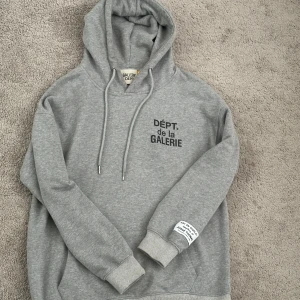 Grå hoodie från Gallery Dept. - Säljer en grå hoodie från Gallery Dept. med svart tryck både fram och bak. Tröjan har huva med snörning, ribbade muddar och en vit patch på ärmen. Perfekt för dig som gillar streetwear och vill sticka ut med coola detaljer. 