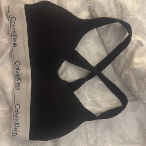 Svart sportig linnetopp Calvin Klein - Svart linnetopp från Calvin Klein med bred, vit resår nertill och logga runtom. Snyggt korsade axelband i ryggen och sportig look. Perfekt för träning eller chill. Mjukt och stretchigt material som sitter skönt mot kroppen.