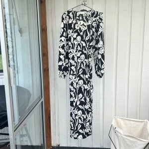 Svartvit omlottklänning med blommor - Snygg lång omlottklänning i svart med stora vita blommor. Klänningen har långa ärmar, knyt i midjan och v-ringning. Perfekt för dig som gillar en stilren men ändå iögonfallande look.
