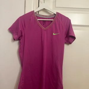 Rosa Nike Pro träningstopp - Nike Pro träningstopp i stark rosa med v-ringning och korta ärmar. Snygg limegrön Nike-logga på bröstet och kontrasterande kant vid halsen. Perfekt för dig som vill ha en sportig och bekväm look på gymmet eller löprundan.