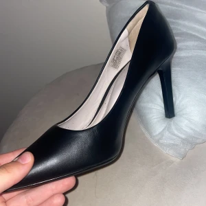 Svarta pumps från Calvin Klein - Klassiska svarta pumps från Calvin Klein med spetsig tå och hög klack. Skorna är i slätt skinn och har en elegant metallplatta med logga baktill på hälen. Perfekta för dig som gillar stilrena och tidlösa skor.