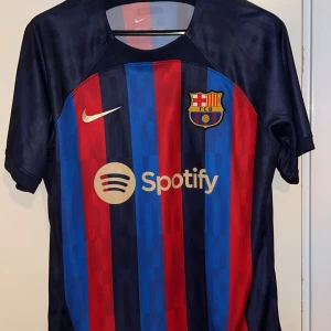 FC Barcelona Nike matchtröja 22/23 - FC Barcelona matchtröja från Nike med Dri-FIT-material. Tröjan har klassiska blå och röda vertikala ränder, klubbmärke på bröstet och Spotify-logga. Kortärmad modell i lätt och ventilerande polyester, perfekt för fotboll eller att bära som supporter.