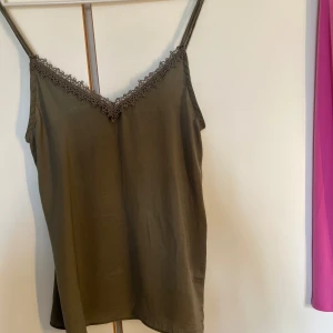 Olivgrön linnetopp med spets från Vila - Supersnygg olivgrön linnetopp från Vila med tunna axelband och v-ringning dekorerad med spetsdetaljer. Lätt och flowy känsla, perfekt för varma dagar eller lager på lager. Enkel att styla till både jeans och kjol.