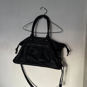 Svart axelväska i skinnlook - Stor svart axelväska med både handtag och lång axelrem. Väskan har en dragkedja framtill och flera fack inuti, perfekt för att få plats med allt du behöver. Tillverkad i skinnimitation med stilrena detaljer och silverfärgade metalldelar. Kostade 1400 och är i äkta läder