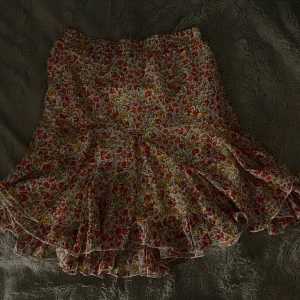 Blommig volangkjol i rött och gult - Supersöt kort kjol med blommigt mönster i rött, gult och grönt på vit botten. Kjolen har volangkant nertill och en luftig, flowy passform med resår i midjan. Perfekt för dig som gillar färg och romantiska vibes.