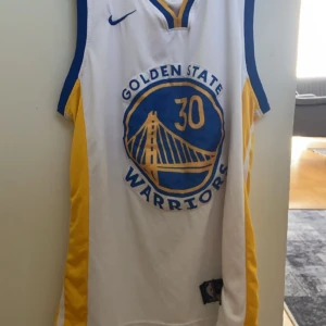 Golden State Warriors Curry linne - Golden State Warriors basketlinne med nummer 30 och Curry på ryggen. Linnet är vitt med blå och gula detaljer, har ärmlös design och är tillverkat i lätt polyester. Perfekt för dig som älskar basket och vill representera Steph Curry på planen eller i vardagen.