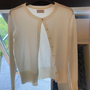 Vit kofta från Holly & Whyte - Supersnygg vit kofta från Holly & Whyte med rund halsringning och guldiga knappar hela vägen framtill. Långärmad och i mjukt, tunt material som känns skönt mot huden. Perfekt att slänga över en topp eller bära stängd för en clean look.