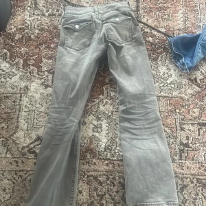 Grå bootcut jeans från Gina Tricot - Säljer ett par grå bootcut jeans från Gina Tricot med snygga fickdetaljer och silverfärgade knappar. Jeansen har låg midja och är tillverkade i klassiskt denimtyg. 