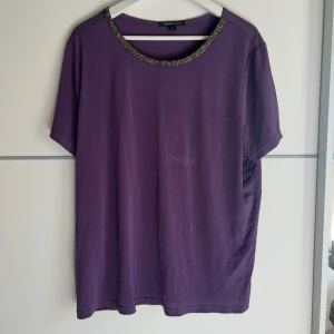 Lila topp med glittrig krage Mywear Woman - Lila topp från Mywear Woman i storlek XL. Rund halsringning med glittrig detalj som ger extra edge. Kortärmad och i mjukt material, perfekt för dig som gillar en enkel men snygg look. Passar dig som vill ha en lite loose fit.