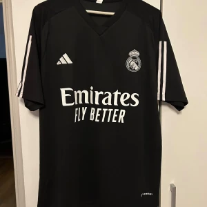 Real Madrid svart fotbollströja Adidas - Snygg svart Real Madrid fotbollströja från Adidas med vita detaljer och klubbmärke på bröstet. Tröjan har korta ärmar, klassiska Adidas-ränder på axlarna och 'Emirates Fly Better' tryckt på framsidan. Tillverkad i lätt och ventilerande material.