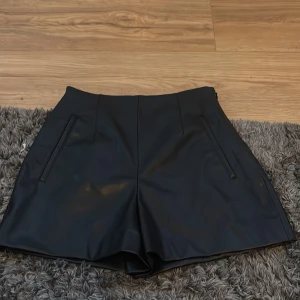 Svarta skinnshorts med fickor - Ser annorlunda ut på❤️ Snygga svarta shorts i skinnimitation med hög midja och raka ben. De har två diskreta fickor framtill och en stilren look som passar perfekt till en trendig outfit. Materialet ger en cool vibe och shortsen är lätta att matcha med olika toppar.