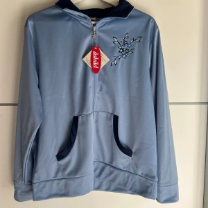 Ljusblå zipjacka med broderi från Åshild - Ljusblå långärmad zipjacka från Åshild med snyggt blommigt broderi på bröstet och mörkblå detaljer vid fickor och krage. Jackan är tillverkad i mjuk polyester och har praktiska fickor framtill. Perfekt för dig som gillar en chill och stilren look.