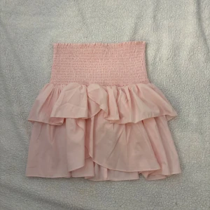 Rosa volangkjol från Neo Noir - Supersöt ljusrosa kortkjol från Neo Noir med smockad midja och dubbla volanger. Kjolen har en flowy känsla och är perfekt för dig som gillar en feminin och trendig stil. Passar perfekt till sneakers eller sandaler för en chill look.