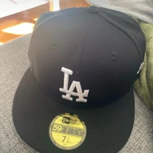 Svart LA 59FIFTY keps från New Era - Snygg svart 59FIFTY keps från New Era med LA-logga broderad i vitt framtill. Aldrig använd och nypris 700. Klassisk flat peak och broderad New Era-logga på sidan. Kepsen är i fast modell och tillverkad i polyester. Perfekt för dig som gillar streetwear och sportig stil.
