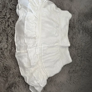 Vit volangkjol med smock i midjan - Supersöt vit kortkjol med volanger och smockad midja. Kjolen har en luftig känsla och passar perfekt till varma dagar. Den är lätt att matcha och ger en fräsch vibe till din outfit.