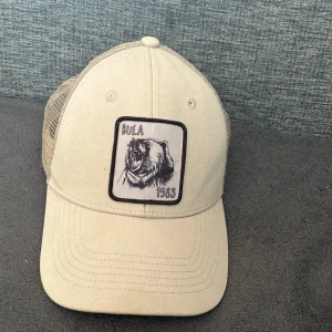 Beige keps från Bula med björnpatch - Snygg beige keps från Bula med mesh-bakstycke för extra ventilation. Framsidan har en cool patch med en ritad björn och texten 'Bula 1983'. Perfekt för dig som gillar streetstyle och vill sticka ut med en unik detalj.