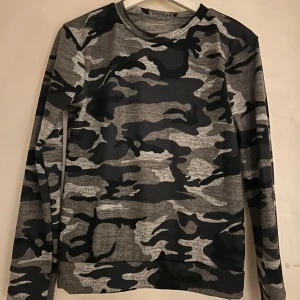 Grå camo långärmad tröja - Snygg långärmad tröja med grått och svart camouflage-mönster. Perfekt för dig som gillar streetstyle och vill sticka ut lite extra. Skön passform och rund halsringning, passar till både jeans och joggers.
